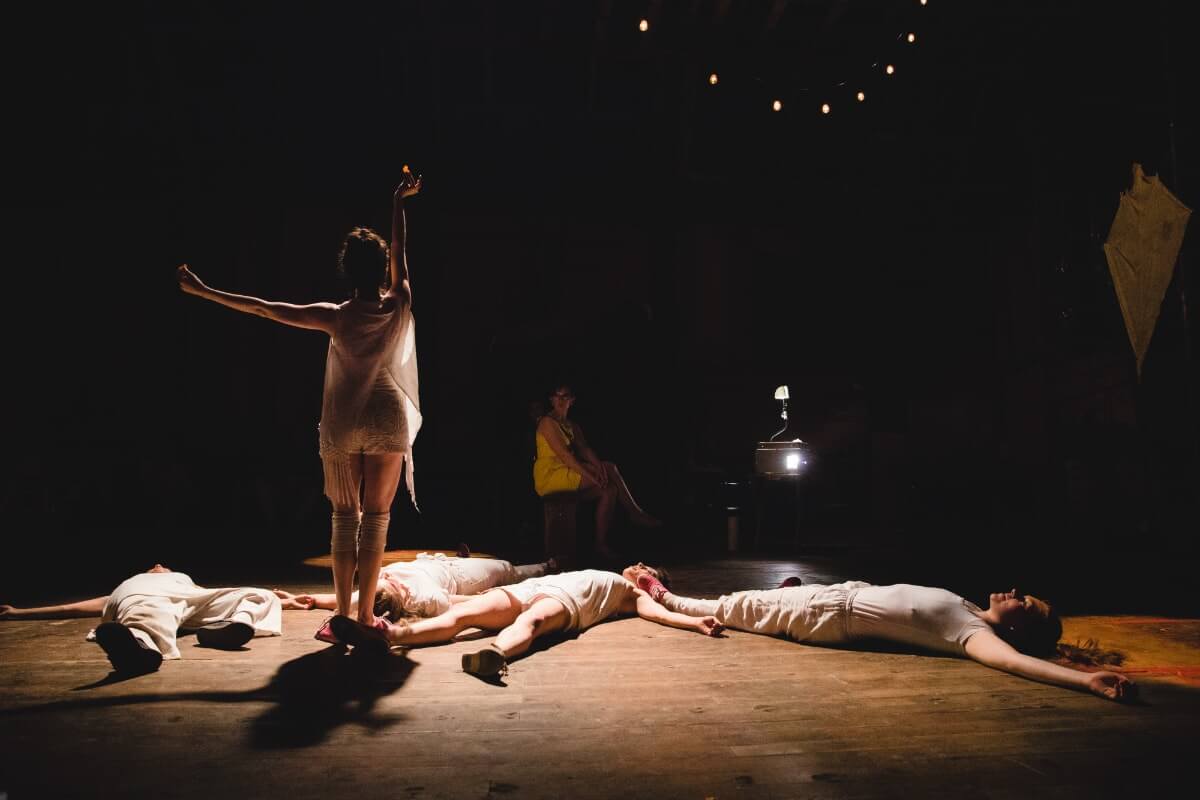A dancer holds a small orange object above her head, while several other dancers lie on their backs on the stage.
