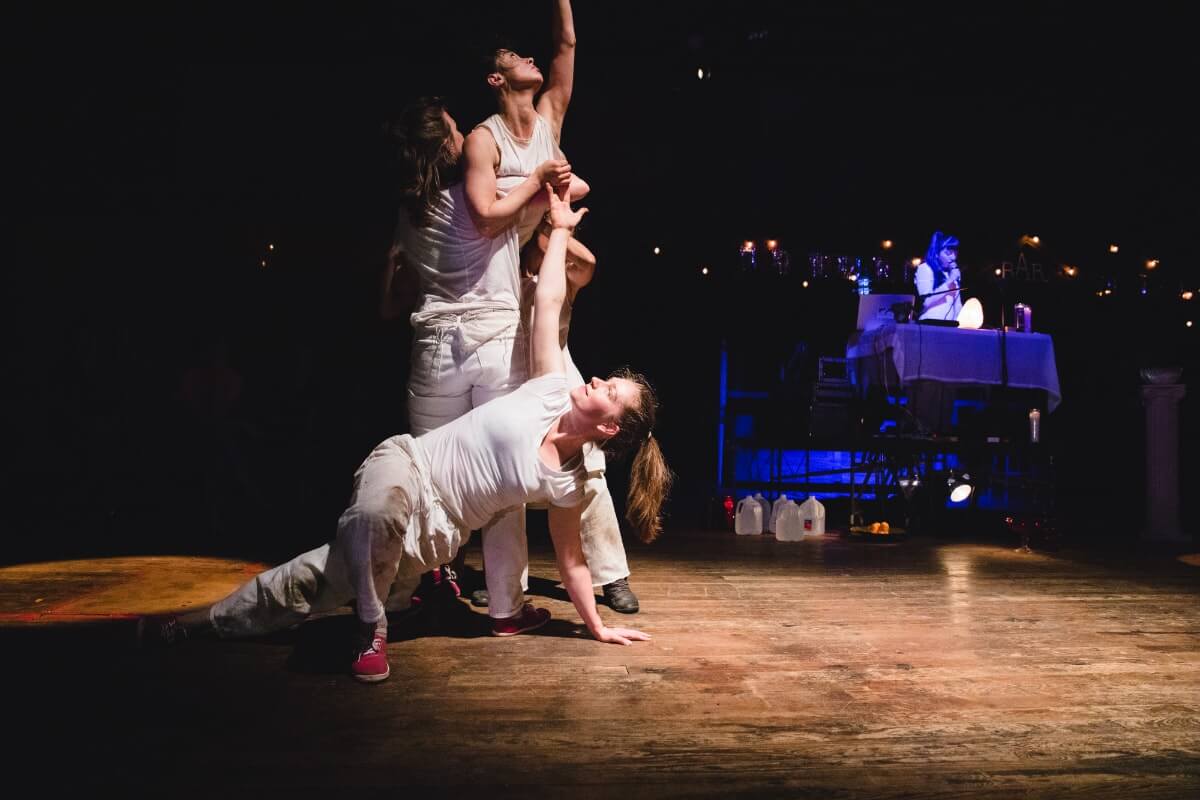 Two dancers lift a third dancer a foot into the air, while a fourth dancer on the ground reaches up as if to offer help.
