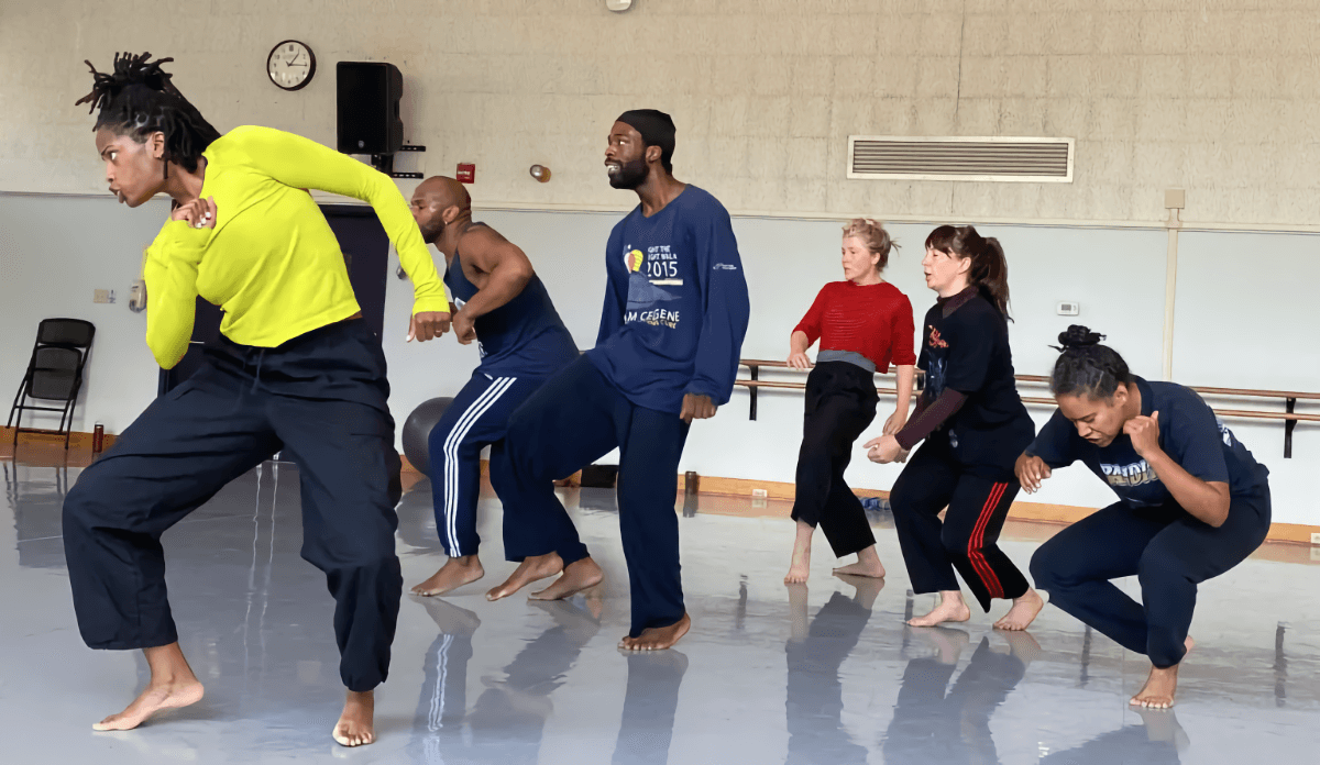 In a large white and beige rehearsal space, Cynthia Oliver and five other dancers of various ethnicities, all barefoot, rehearse.