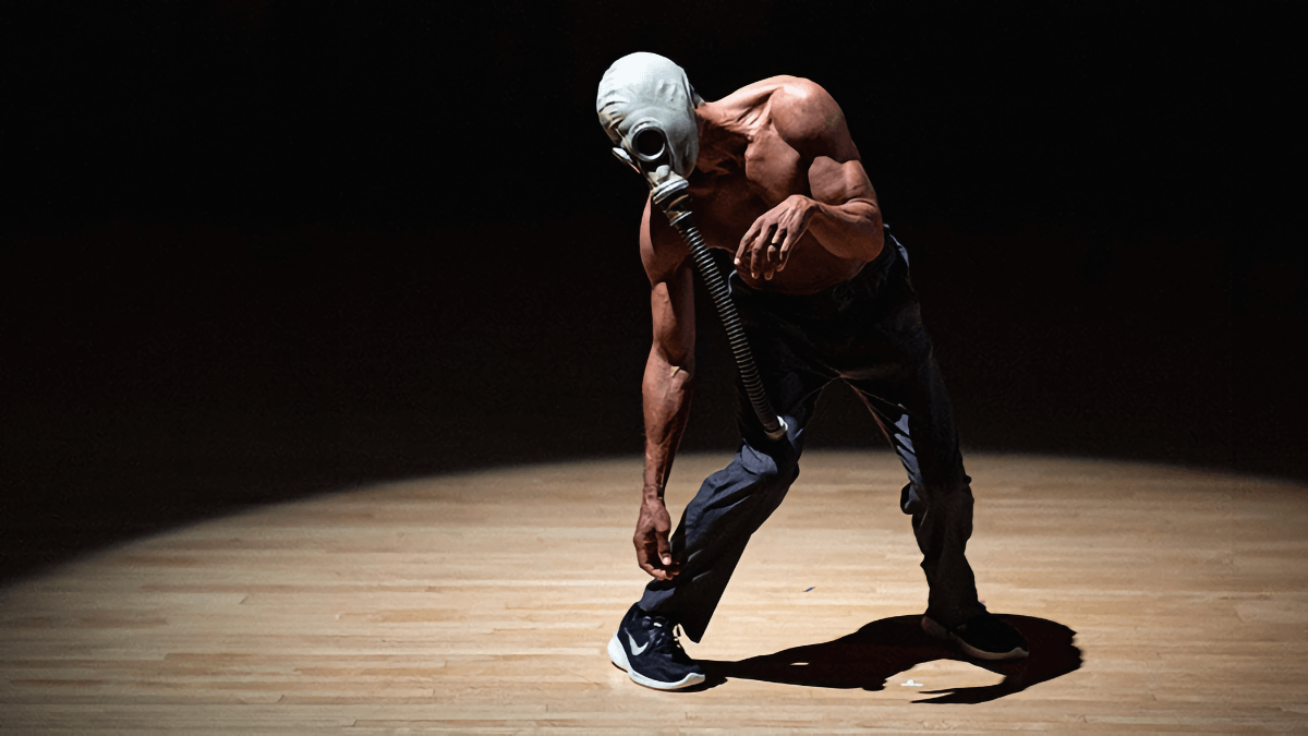 Pictured: David Roussève. Photo: Rachel Keane. A man in a stark pool of light leans heavily to the left, with one arm dangling low to the ground. He is wearing an old gas mask, and the breathing tube also dangles from his face, disconnected from any source. His upper body is bare, revealing his medium brown skin and the musculature of his torso; on his lower body, he wears dark pants and black running shoes with white swooshes.