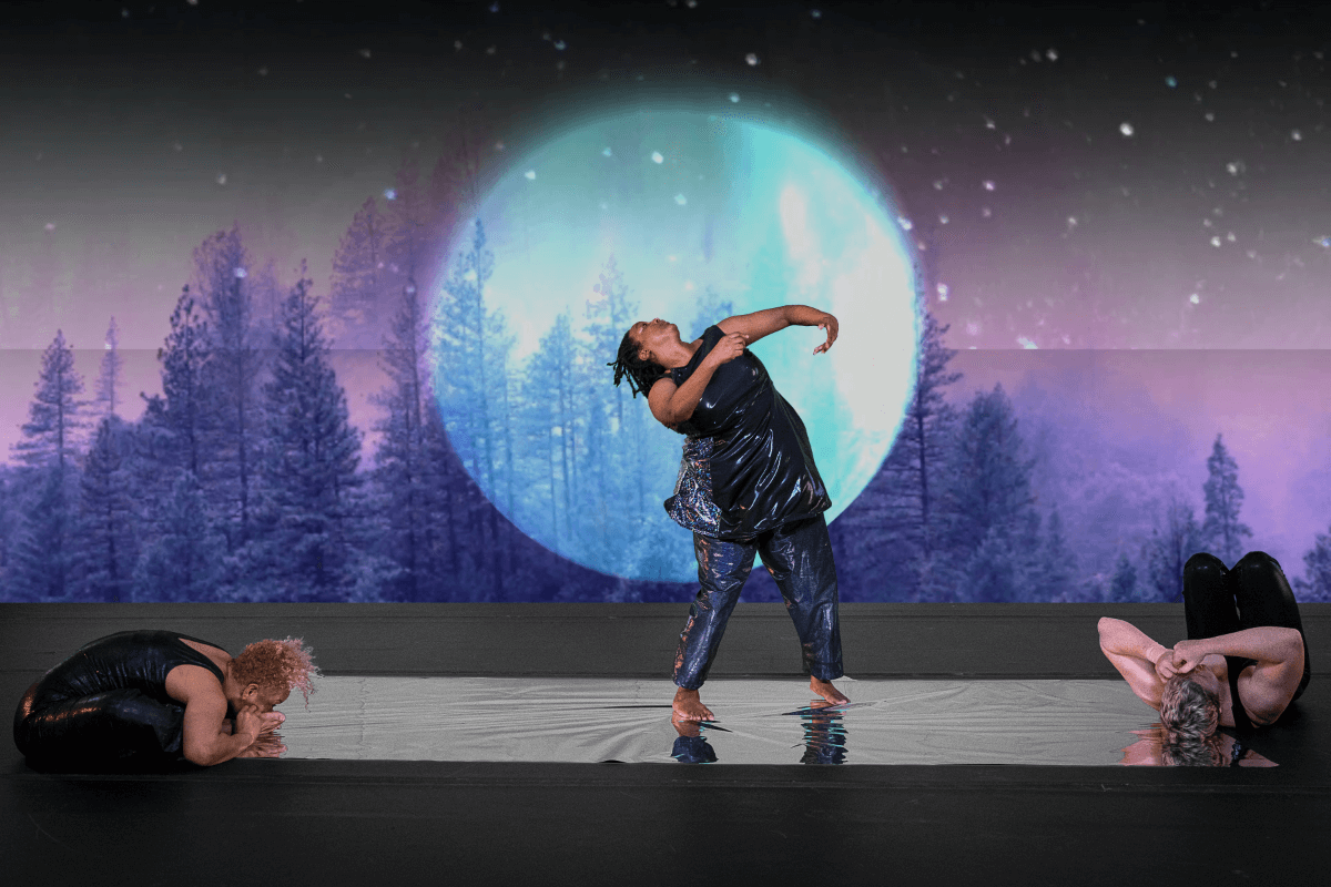 Kayla Hamilton stands on a reflective strip of silver, chin lifted as she arches her torso backwards, arms curved in front of her chest. Alice Sheppard sits on one end of the silver floor, torso folded into her own lap. Laurel Lawson lays on their back at the other end of the silver. Alice is a multiracial Black woman with short curly blonde hair; Kayla is a Black woman with shoulder length locs; Laurel is a white person with cropped hair. All three dancers wear dark shimmery bodysuits, a detailed video projection of a huge luminescent moon and a dense forest drenched in hues of cool blues and rich purples fills the background.