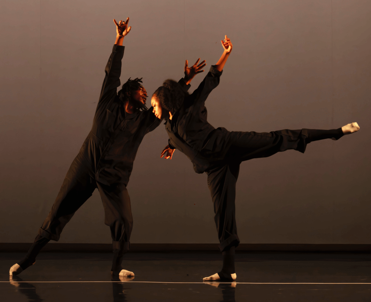 Latrea Rembert and Chandler Bingham extend their limbs on a dimly lit stage as they face each other. Latrea gestures his arms above him, his eyes following their path as Chandler glances downward, her leg extended behind her at ninety degrees. They wear matching black jumpsuits and legwarmers, accented with crisp white socks on their pointed feet.