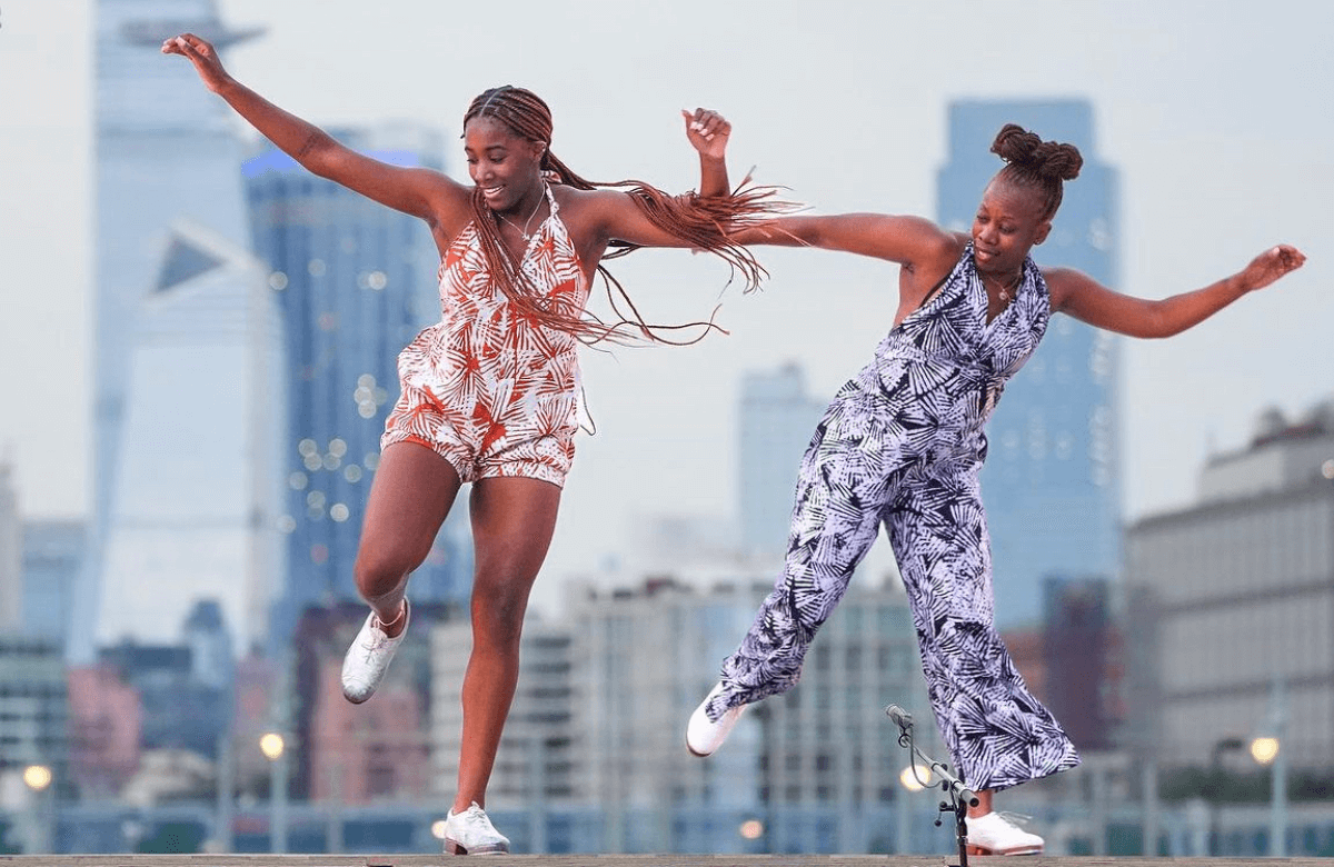 Two Black women are mid-movement tap dancing with raised arms and raised right foot. The dancer on the right wears a blue and white patterned jumpsuit, and the dancer on the left wears an organ and short white jumpsuit.