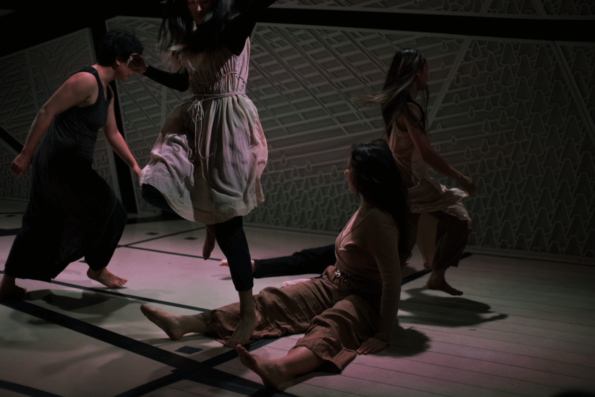 A short-haired medium-skinned Asian woman wearing a black tank and pants, a medium-skinned Asian woman with long hair and blonde tips wearing a light pink dress over a black turtle neck and navy cropped pants, and a deep medium Asian woman wearing a nude spaghetti strapped dress over cropped nude pants, run and jump over the legs of two performers sitting with their legs in a V. The seated performed at the forefront is a light-skinned Asian women with dark long hair and nude colored shirt and pants. The one seated behind is wearing black and only her feet are visible.