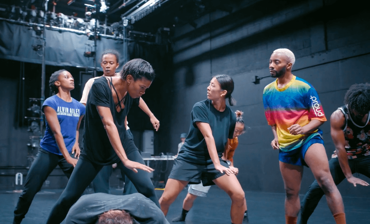 A group of eight dancers of various ethnicities rehearse in a large, empty, brightly lit performance space with the floor, ceiling, and walls painted black.