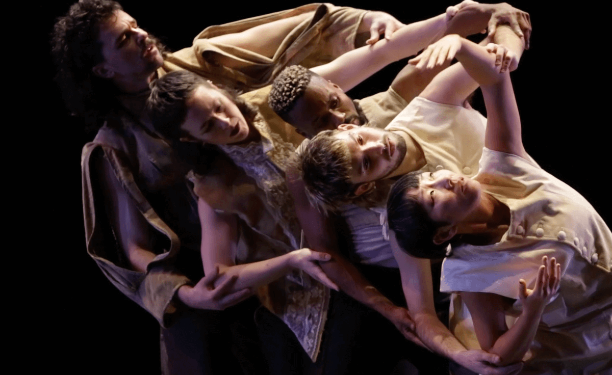 On a black box stage 5 performers are linked together in a human chain with their hand in contact with the performer in front's elbows. From back to front a white non-binary person with a dark mustache, a white non binary person with a dark mullet, a black man with frosted bleached hair, a white non binary person with bleached tip mullet and a asian woman with a short blunt dark haircut all make a stream with their physical connection as the person in front reaches their arms up in a delicate extended gesture.