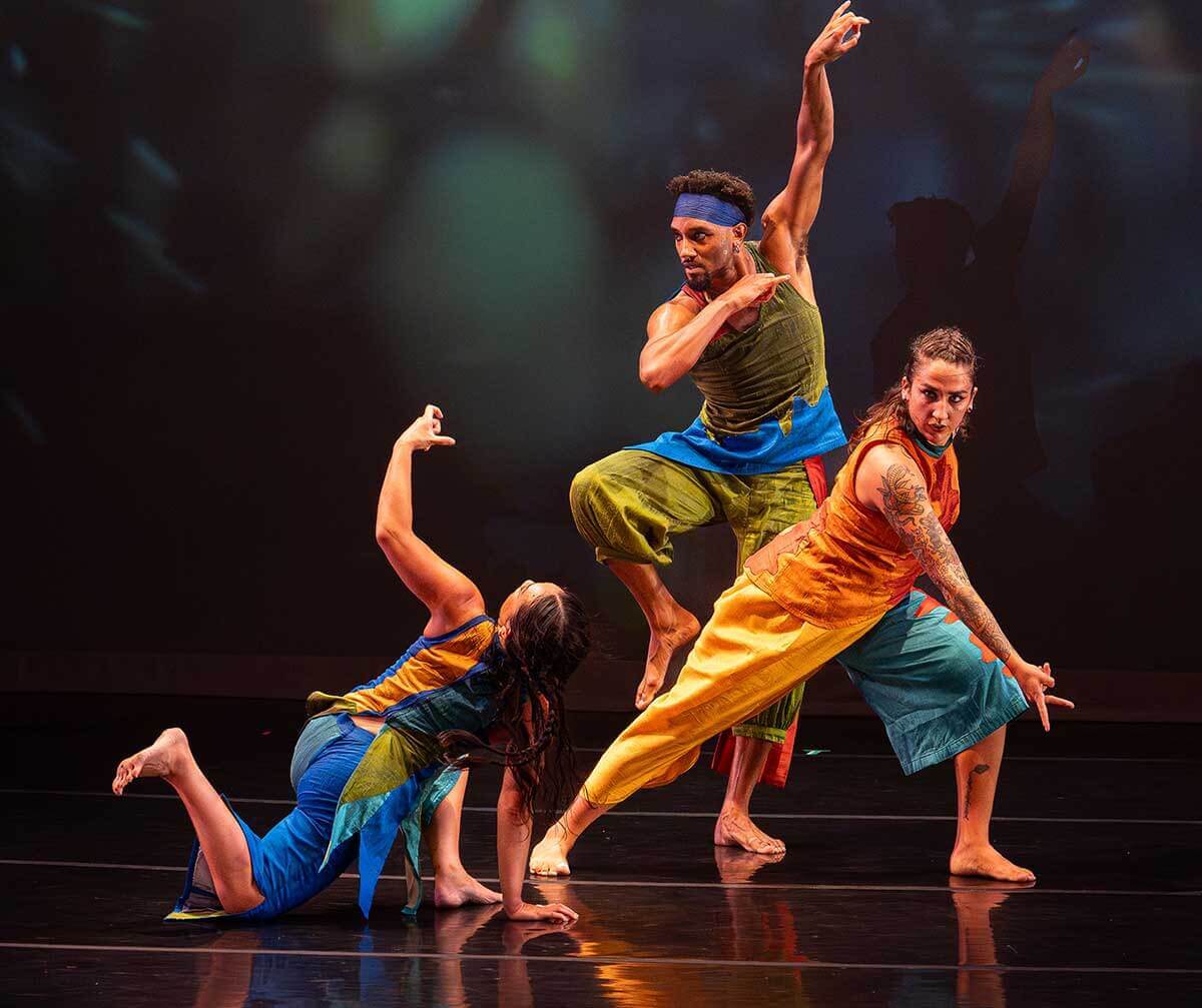 A trio of dancers of varying skin tones wearing jewel-toned multicolored tunics and pants hold a dynamic pose, bodies overlapping and almost intertwined. The Asian American woman kneels in a lunge with one foot and one arm extended into the air, toes energetically extended and fingers crooked in a mudra (a symbolic gesture rooted in Hindu practice). The African American man plants one foot on the ground, the other leg raised at the knee and one arm extended overhead, fingers delicately circled in a mudra. The Palestinian American woman lunges to one side, her back leg and torso and head in sharp alignment, her front arm and fingers casting a mudra down towards the ground.