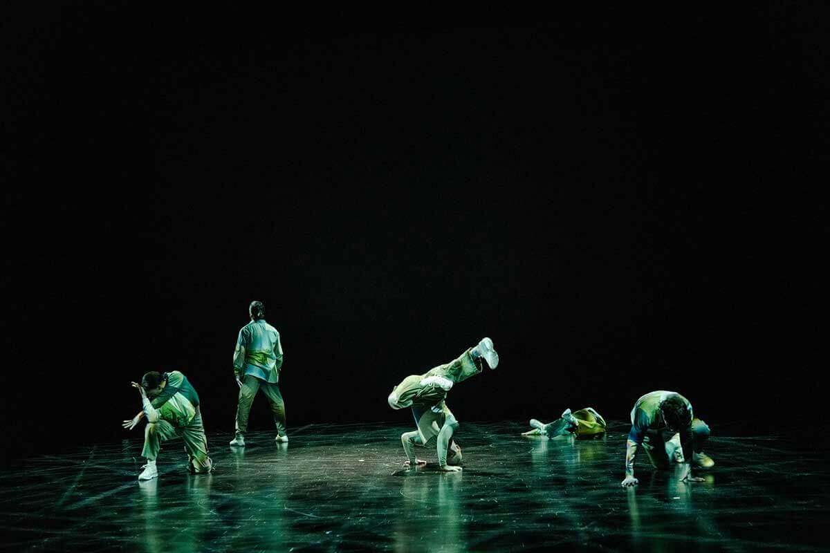 Five dancers on a proscenium stage. There is a male dancer in the middle that is surrounded by the other four and he is balanced on both arms with legs in a figure-4 position in the air—his body shaped like an axe or hammer. All dancers are wearing shades of green and sneakers.