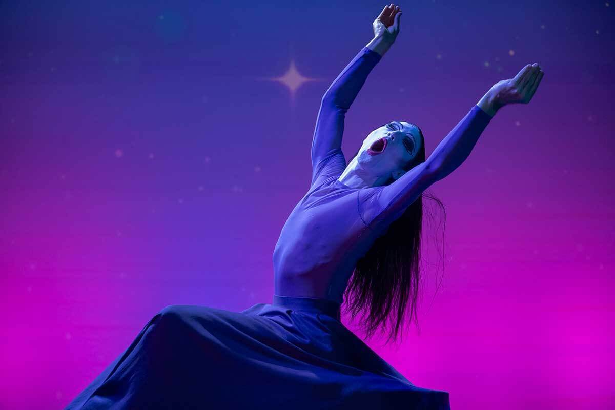 An individual with a painted face and long dark wig gestures with bent elbows and sculpted cupped hands overhead. 