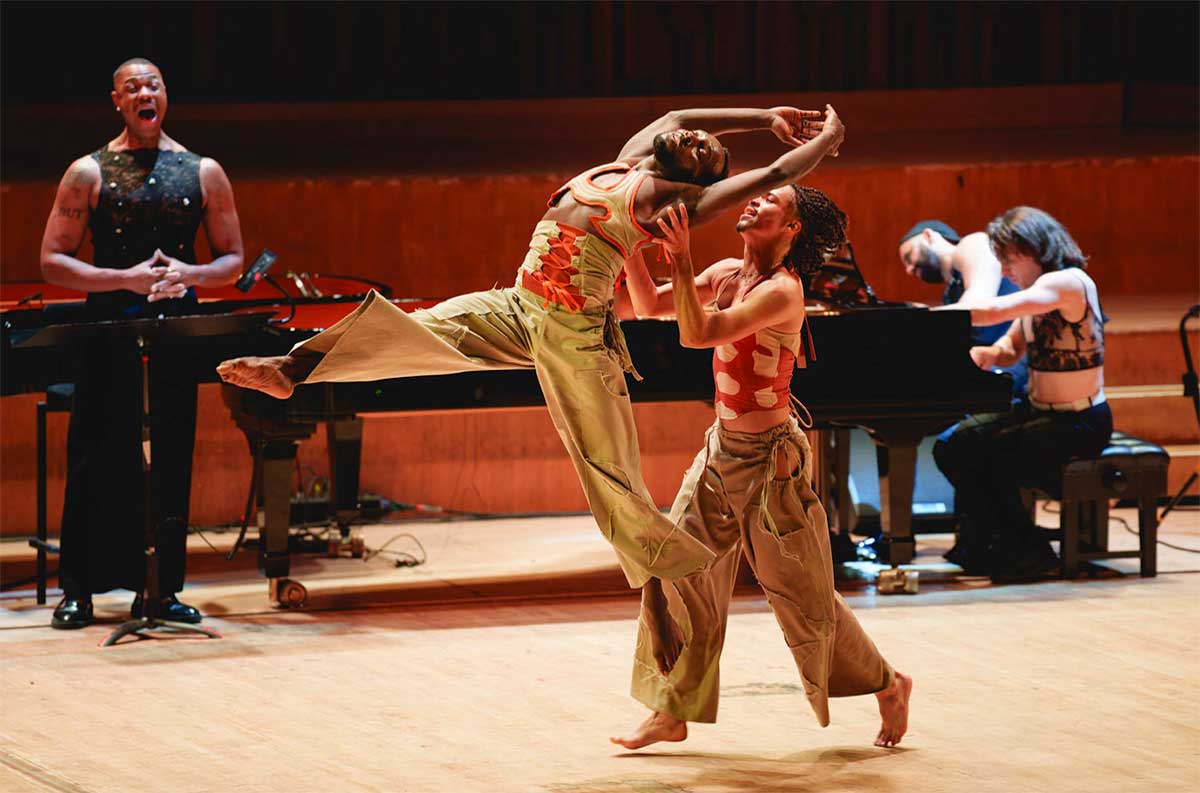 Description: A performer of color joyfully lifts another as he reaches above his head with a vocalist and pianists in the background // Costume + Makeup Edo Tastic (KMC Creative Director)