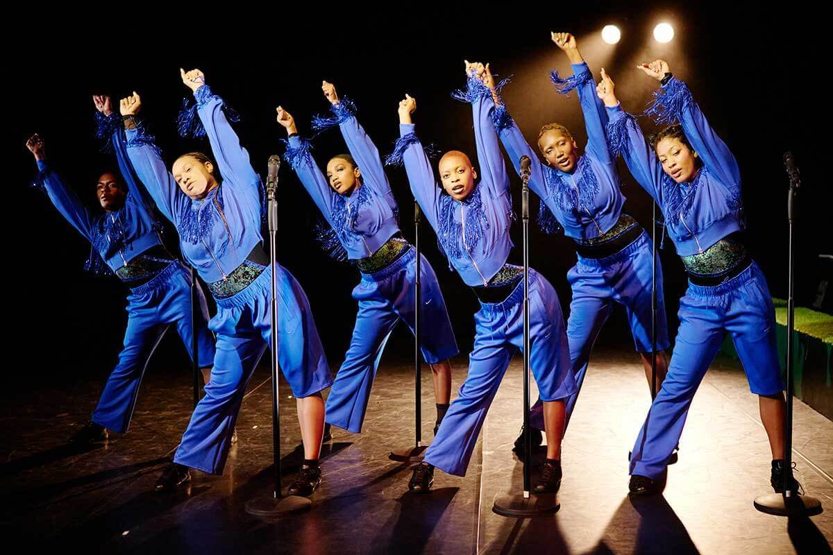 6 Black femmes in blue track suits with sparkling fringe across the chests and wrists stand in a staggered line before 6 microphones. Their hips are popped to one side and both arms are raised above their heads and tilted at a diagonal. 