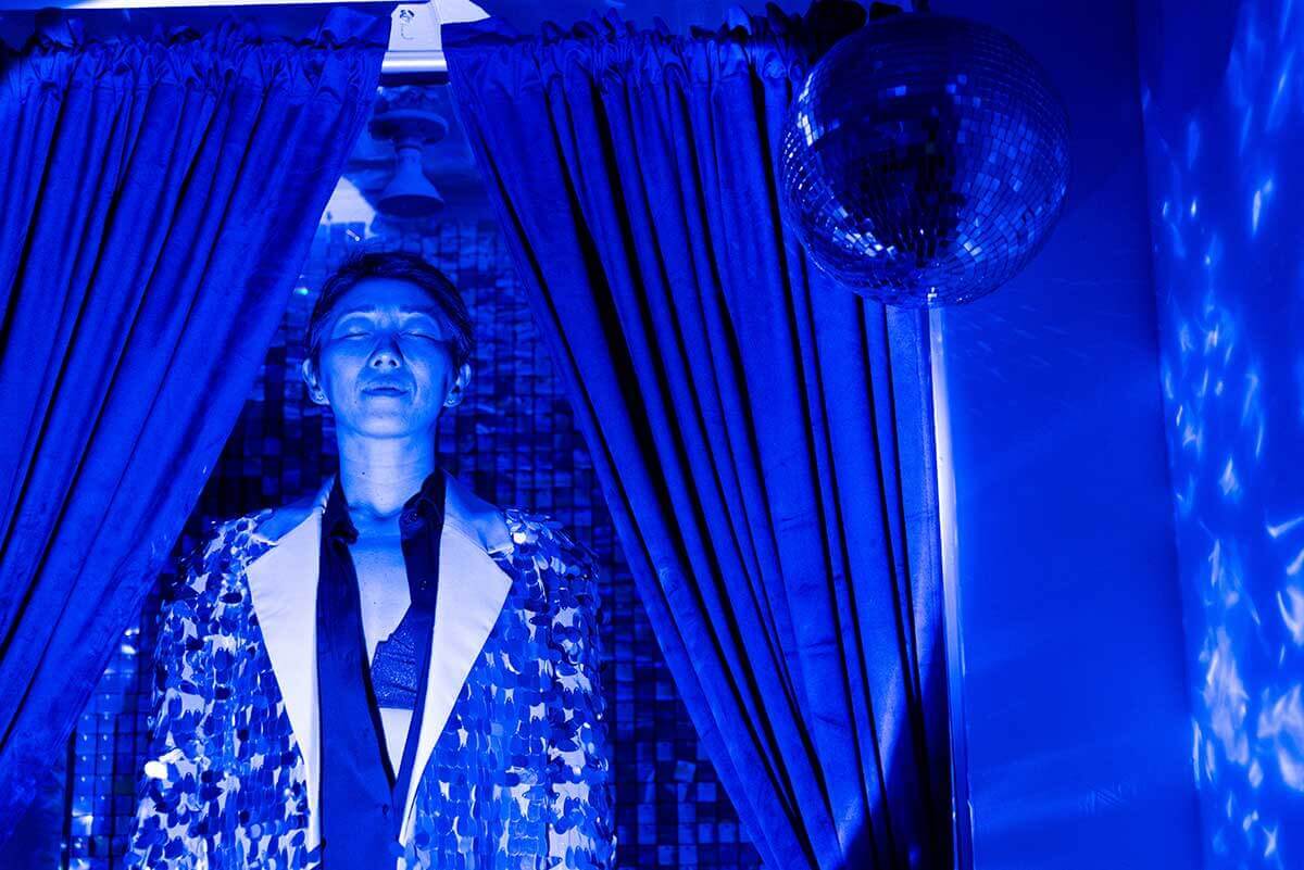 A light-medium skinned Korean-American woman from the waist up, standing upright with arms at her side with her eyes closed in the blue hue of DIY lighting. Two curtains frame her, shiney tiles hang behind her, and a disco ball hangs from the ceiling in front of her to the right casting light on the wall. She is wearing an oversized sequin jacket over a black, silky button up blouse that is partially buttoned revealing a sparkly black bra.