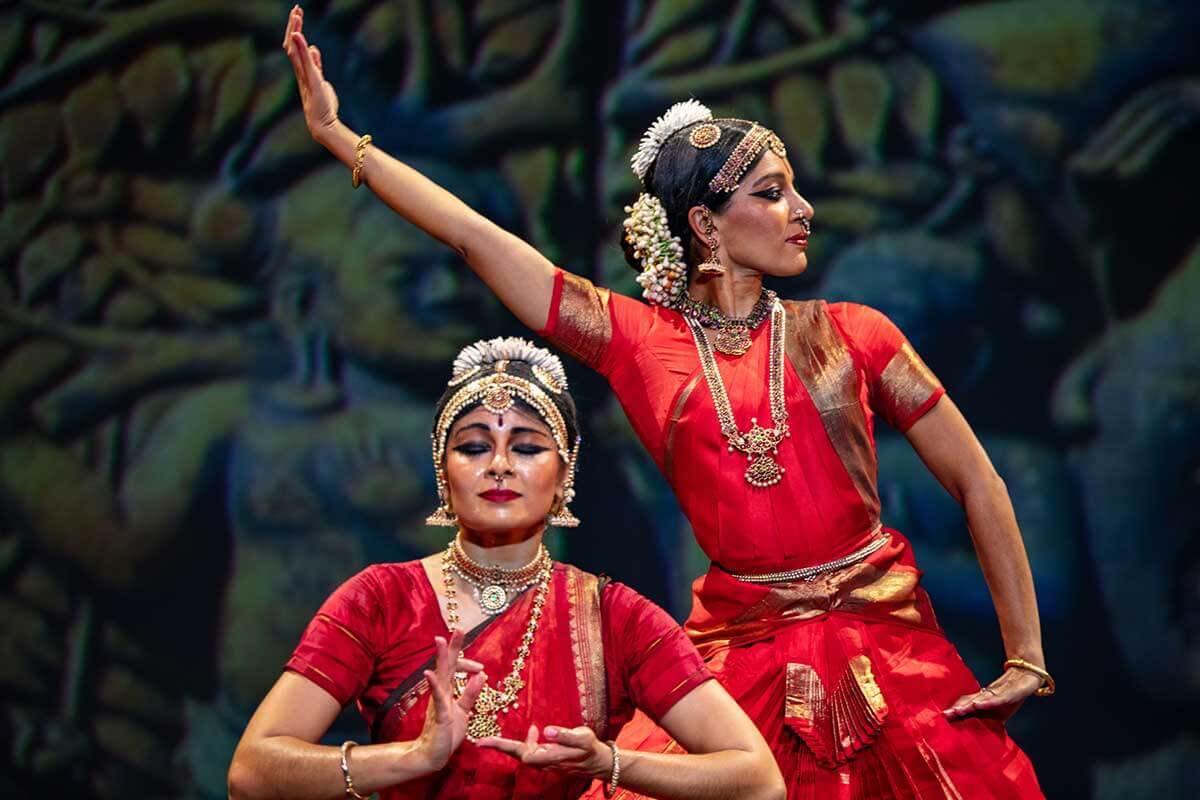 Two women perform a classical Indian dance in traditional Bharatanatyam attire, featuring vibrant red silk costumes with gold borders, ornate jewelry, and fresh flower garlands in their hair. One dancer holds a graceful hand mudra while the other extends her arm dramatically, both set against a dim, artistic background that enhances the cultural richness of the scene.