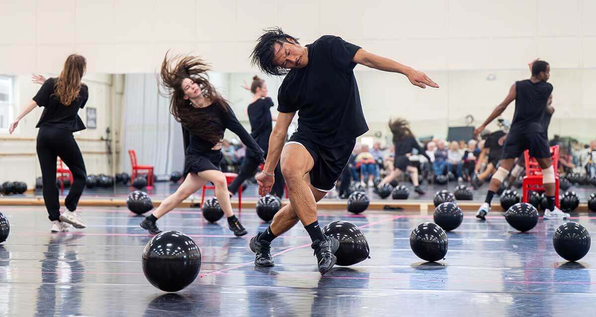 A group of dancers is mid-movement in a spacious studio filled with glossy black balloons scattered across the floor. At the center, a medium-skinned dancer with straight, shoulder-length dark hair moves with a deep side lean, arms extended, wearing a loose black shirt, black shorts, and athletic shoes. Behind and around him, several other dancers (of varying skin tones including light-skinned, and dark-skinned) are captured in dynamic motion, their hair and limbs sweeping through space. All are dressed in black dance attire. A mirrored wall reflects the dancers and a seated audience watching from the back of the room.