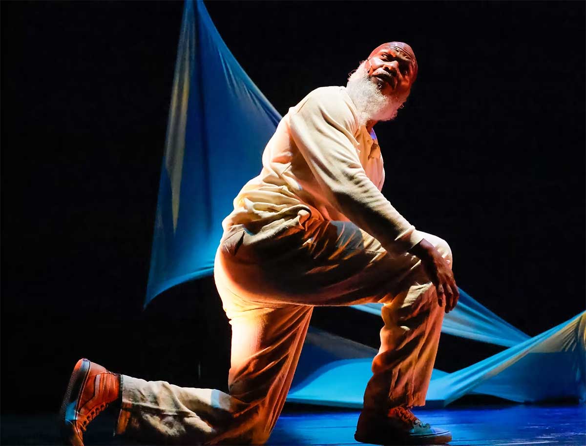 A male performer with a white beard and head covering kneels on stage in dramatic lighting, wearing cream-colored clothing and colorful sneakers. The background features flowing blue and white fabric, adding movement and depth to the theatrical scene.