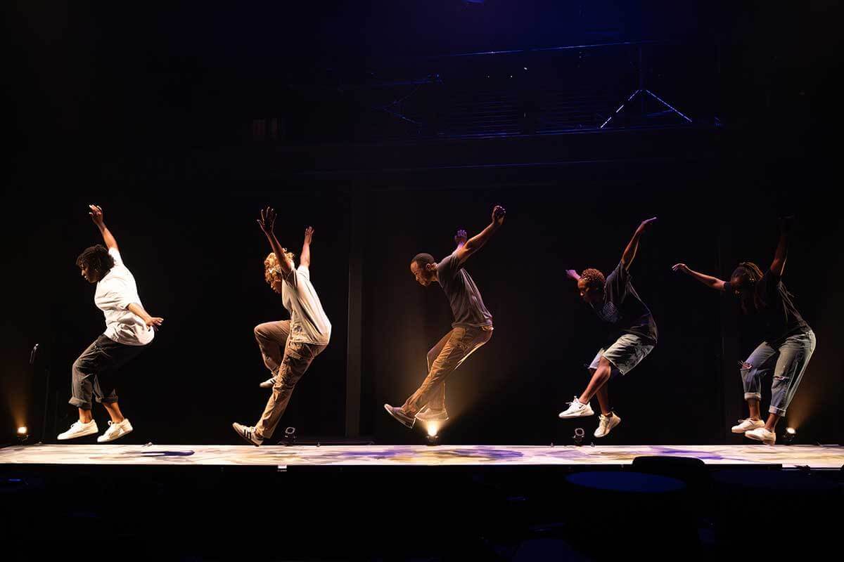 Six dancers with a range of brown skin tones — five female-identifying and one male-identifying — perform a dynamic group sand dance. Their bodies are lifted mid-air, arms extended high as they sweep sand across the floor, creating a striking rhythm of movement, texture, and energy.