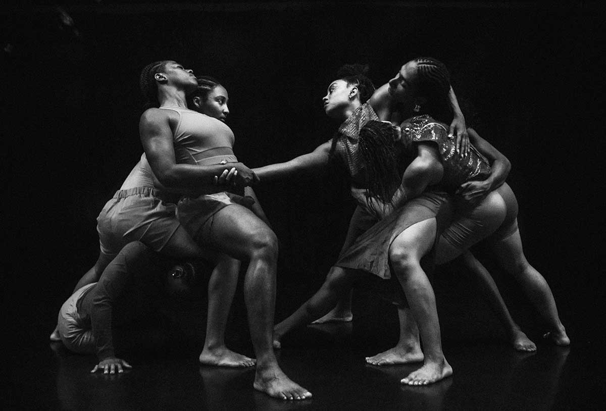 Six female black dancers in various positions of embrace separated into two groups of three with the groups connected by the arm of one dancer. 