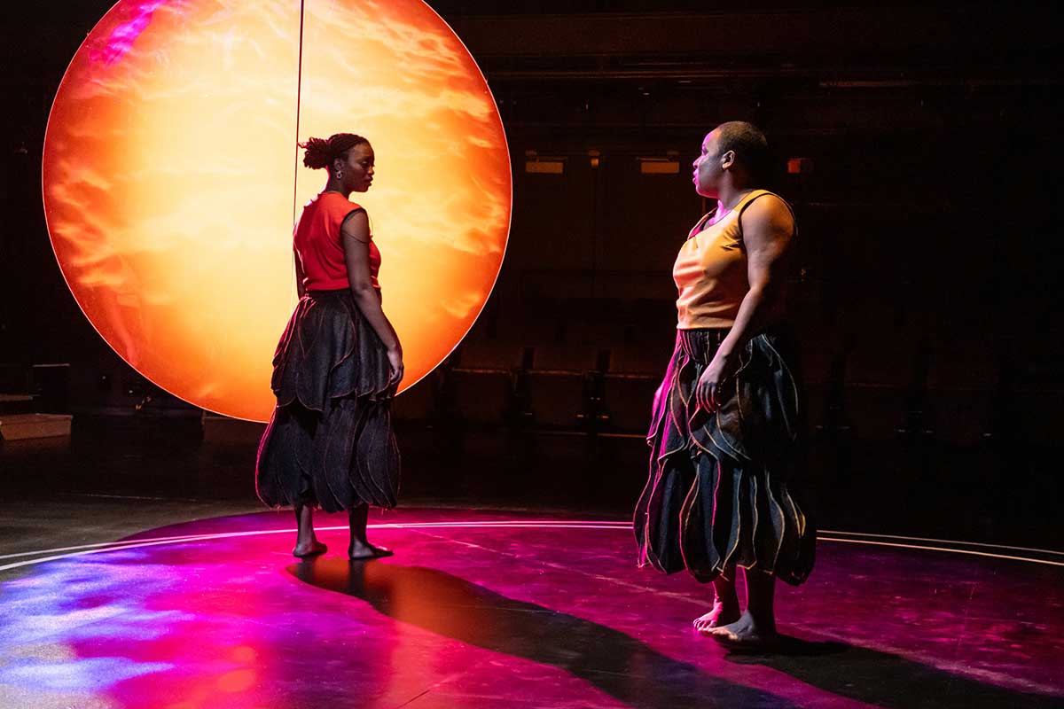 A medium-skinned woman stands in front of a large orange and yellow glowing circle. She faces backwards towards a medium-skinned woman in profile. The first woman wears an orange tank top and flowing, layered black pants, and the second woman wears a yellow tank top and the same pants. They are both barefoot, and the black floor is illuminated by pink light.