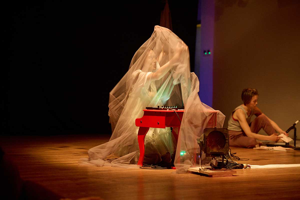 An Asian woman with dark hair sitting at a red toy piano mid-movement dancing. She is moving under apricot-colored tulle, arms over her head, palms touching the tulle, looking up. A small midi controller sits on top of the toy piano. Between the legs of the toy piano is a green light coming from equipment. In front of the toy piano is a lightShe is wearing a white short-sleeved blouse with a light green dress with ruffles at the bottom. In the background to the right is another short-haired performer sitting on the floor. She is wearing a gray tank and brown pants holding paper under a microphone.