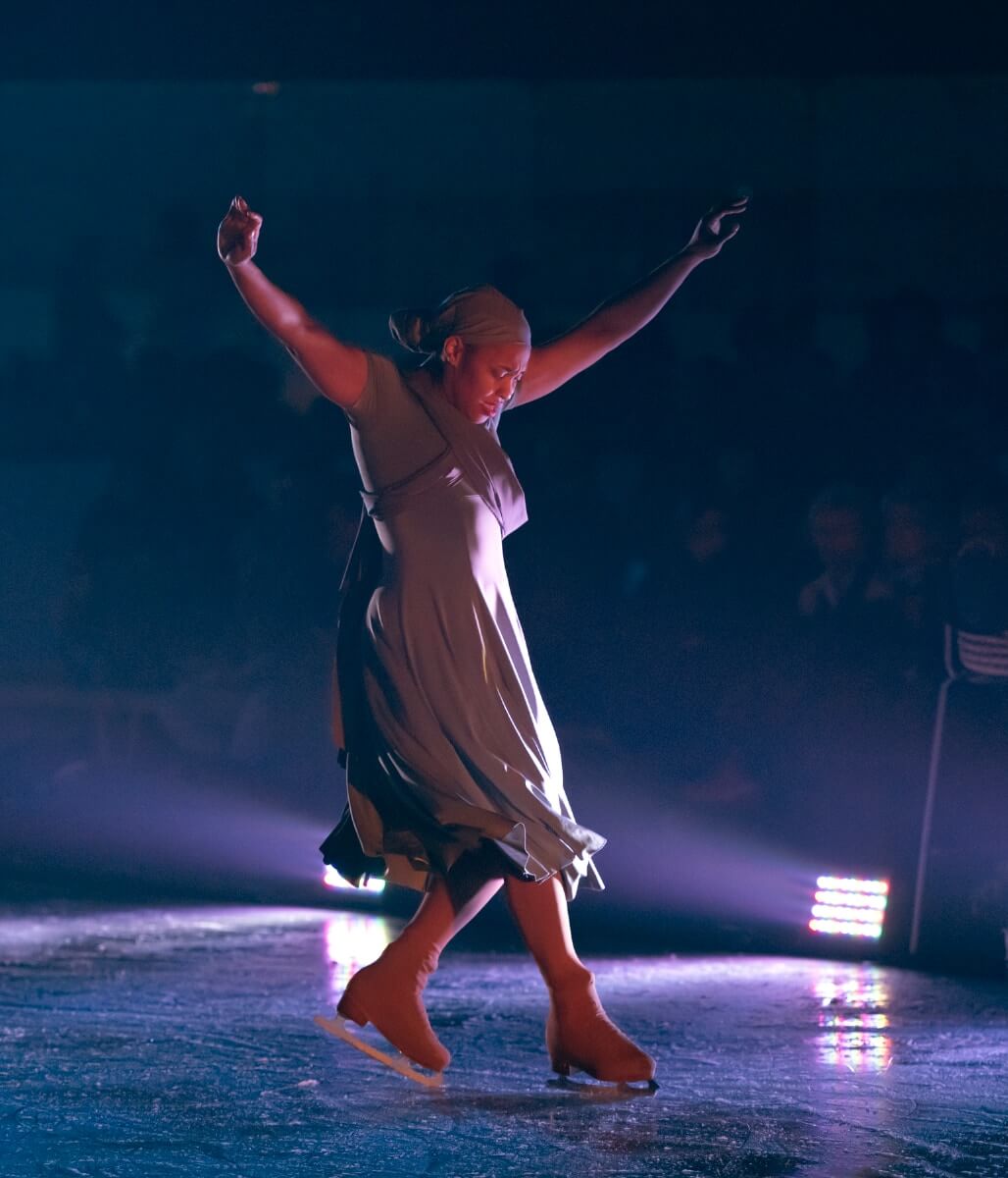 A Black woman on the ice in figure skates mid-movement balancing on the toe pick of her left skate while extending her right foot forward with both arms extending over head. She is wearing a gray dress with a full skirt and matching gray headwrap.