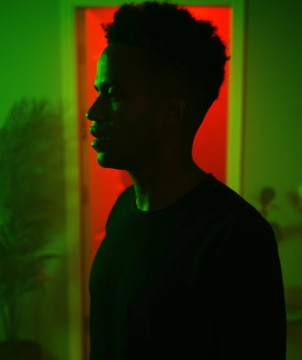 A Black man with a curly flat top dressed in all black is profiled to his right as he stands in a shadowy green room. He stares straight with a troubled look 10 feet in front of an open door where haze and a bright red light emanate from.