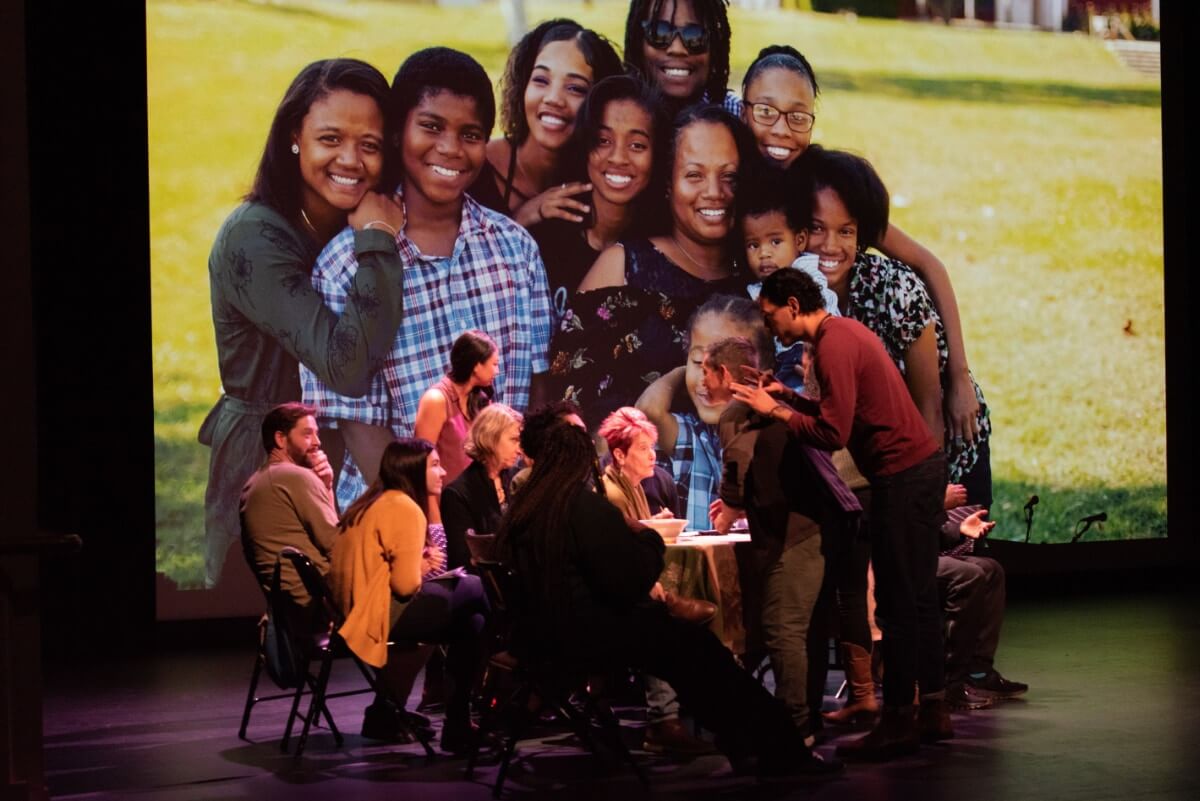 A group of people sit at a table looking up at three performers who are leaning over them: A brown-skinned woman, an older white man, and a tall brown-skinned man. A projection of a smiling African American family is on the backdrop behind the table.