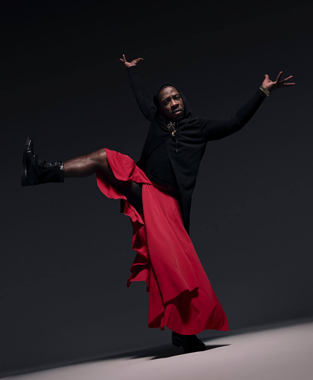 Black man dancing with one leg and both arms raised. He is wearing a long red skirt and a black long-sleeve shirt.