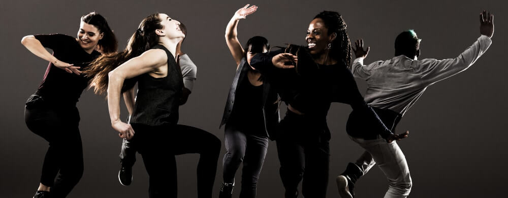 Five dancers is various positions, arms and legs in the air in front of a gray background.