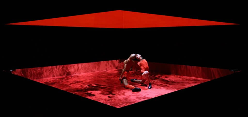 2 men of color lunge towards each other, heads almost touching; feet almost touching. They are both dressed in red tight-fitted athletic/wrestling clothing while inside of a red boxed space. The floor displays a red moving lake, while the interior of the box is engulfed in a striking deep red color. The outside of the boxed ring is black with the middle cut out which functions as the viewing point into the red box.