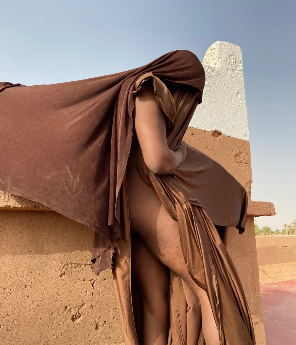A brown skinned body is draped in brown materials. The legs are exposed and the garment is slightly wind blown. 