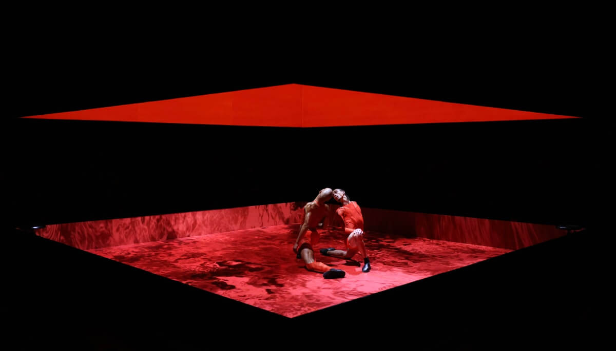 2 men of color lunge towards each other, heads almost touching; feet almost touching. They are both dressed in red tight-fitted athletic/wrestling clothing while inside of a red boxed space. The floor displays a red moving lake, while the interior of the box is engulfed in a striking deep red color. The outside of the boxed ring is black with the middle cut out which functions as the viewing point into the red box.