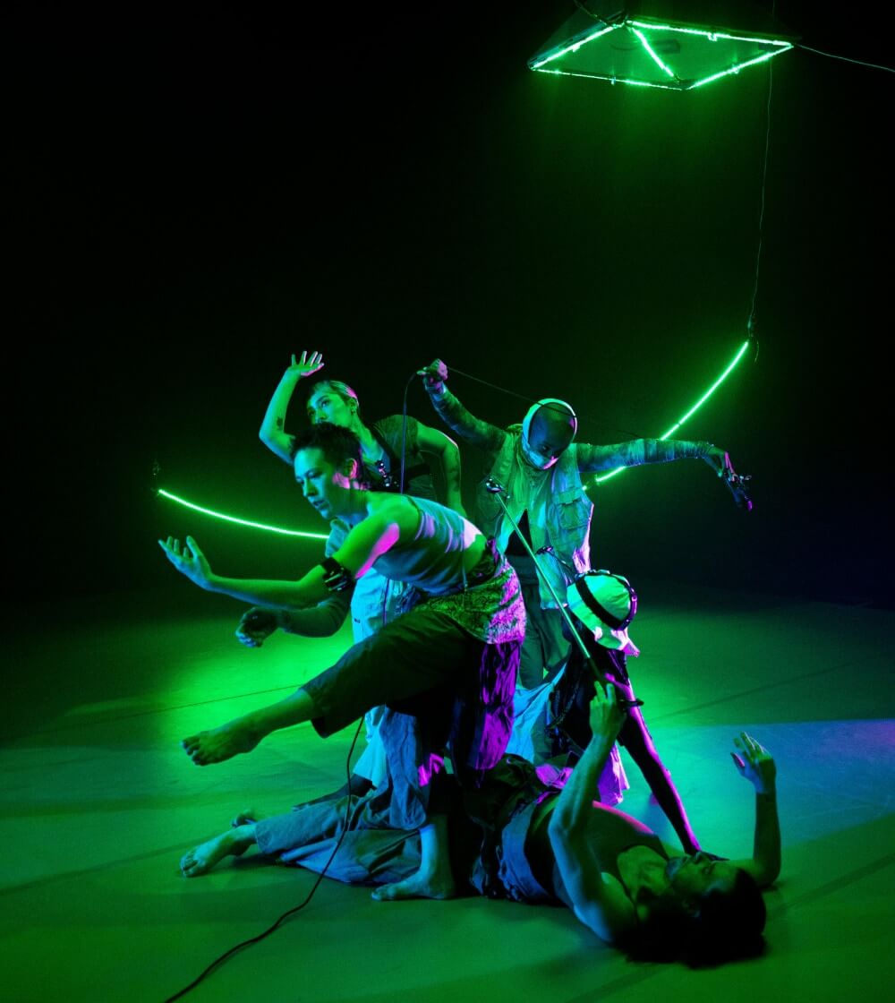 Four dancers are in a clump in the foreground with various body shapes. There are neon green objects lights on the floor behind them and floating above their heads.