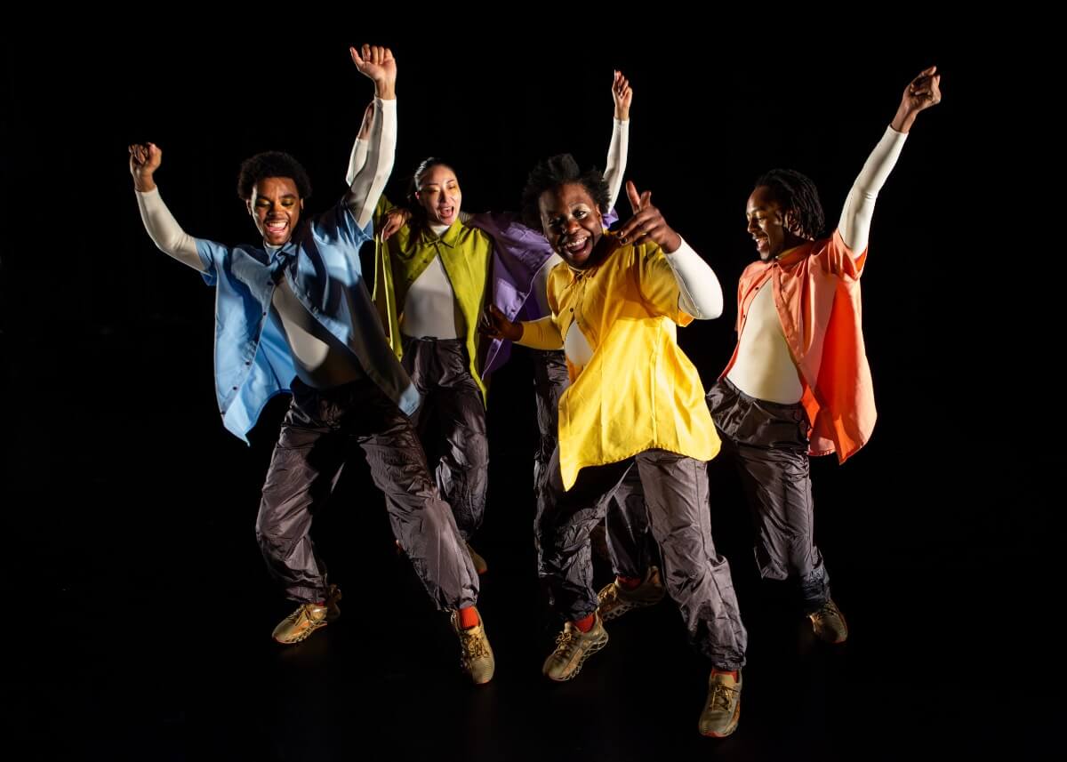 Four black dancers with their arms raised are in front of a black background. They are wearing brightly colored shirts, black pants, and brown shoes.