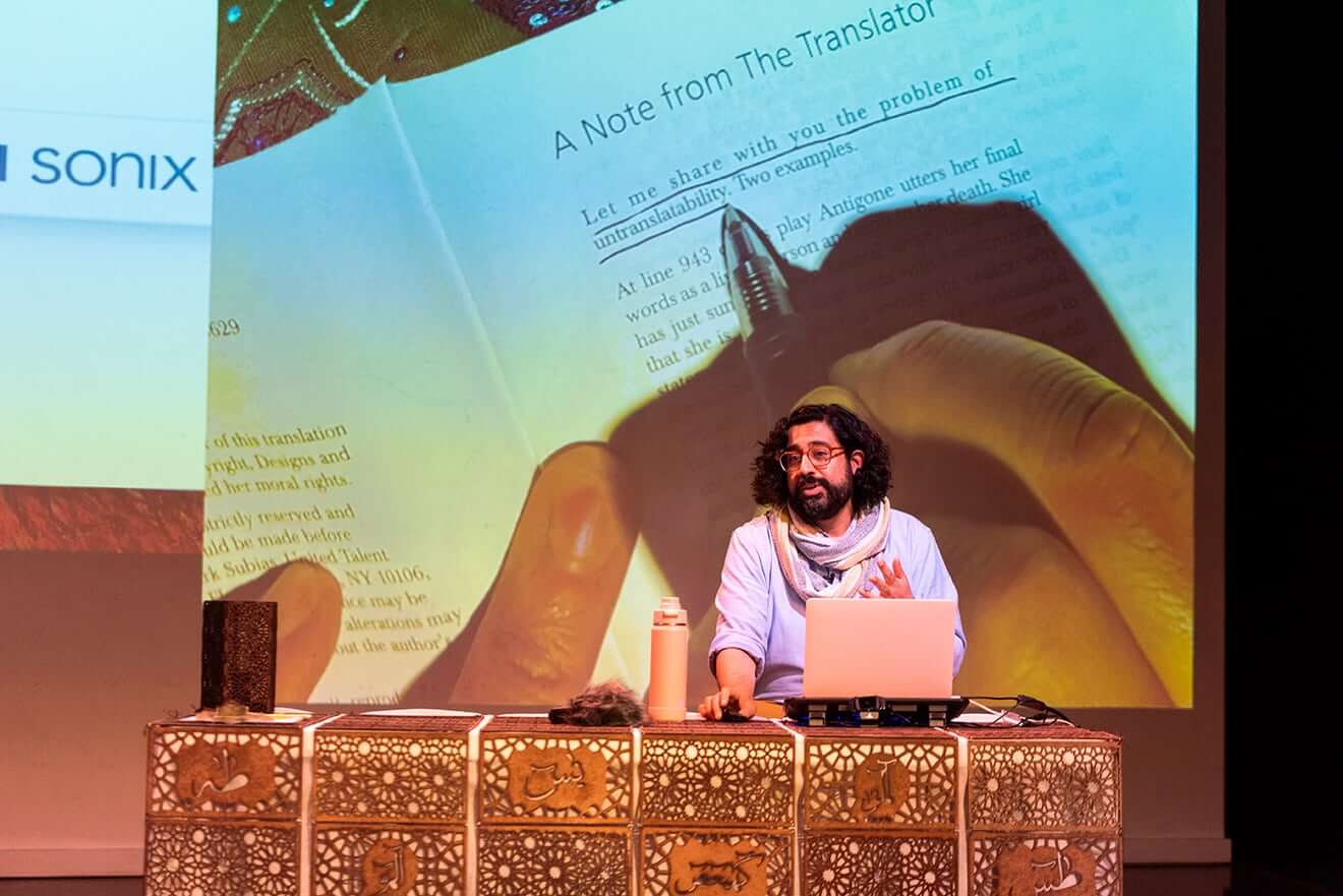 Artist Adil Mansoor sits at an intricately carved wooden table with a pink laptop and pink water bottle. Adil is of Pakistani heritage with curly black hair and a beard. He is wearing glasses, a blue button-down shirt, and an infinity scarf. He is addressing the audience with a projection behind him of a page from a book. The quote “Let me share with you the problem of untranslatability” is underlined in the photo.