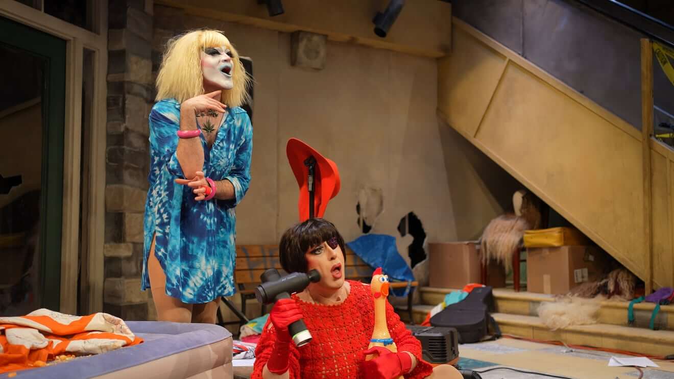 Carla Rossi, a whiteface drag clown with blonde shoulder-length hair, a cannabis pendant necklace, and a tie-dyed blue dress, stands over Pepper Pepper, a white drag queen with a black bob, a red crocheted dress, and a bedezzaled eyepatch. Pepper holds a rubber chicken and pulverizes her face with a massage gun while Carla lipsyncs above her with wide, painted-blue lips, both of them next to an inflatable air mattress on an apocalyptic theatrical set.