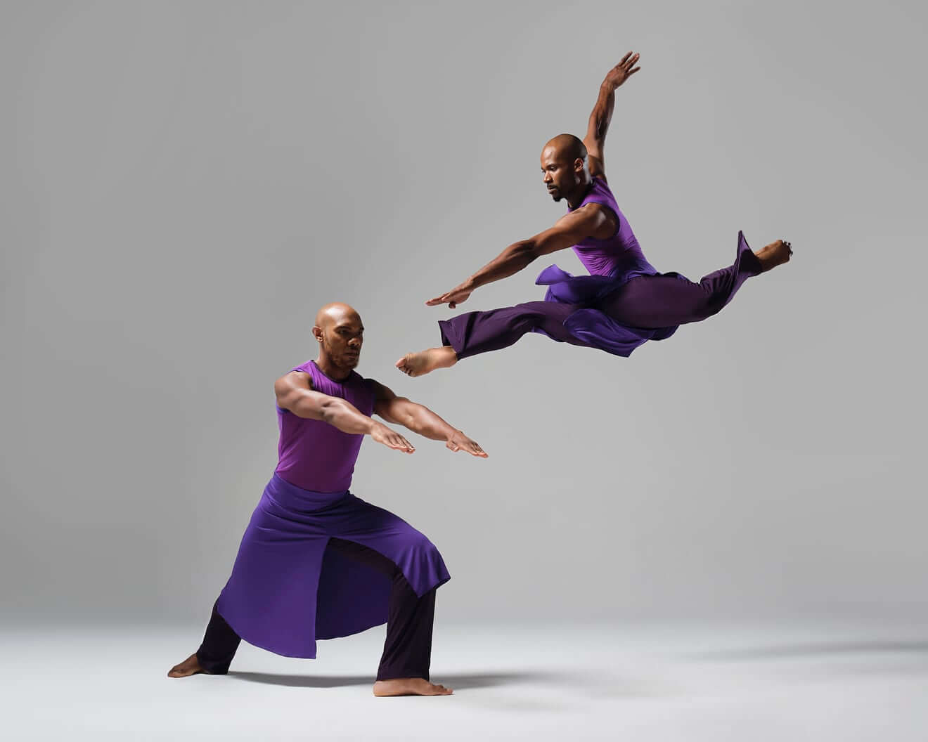 Two Black men dance against a white background, wearing royal purple costumes with sleeveless tops, pants, and flowing skirts with a slit in the front. One dancer lunges forward on one leg, the other extending behind and both arms reaching straight forward. The other artist leaps high in the air, legs slightly bent in a split.