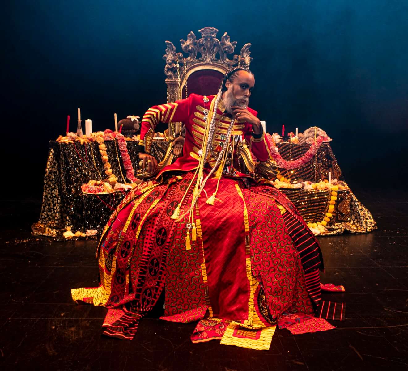 Kaneza Schaal as King Leopold II sits on a throne with her left hand lifted to her chin, left elbow leaning on the armrest, and right hand on the right armrest, elbow lifted, looking at the viewer. She wears a white beard, hair in two long white braids, and a red and gold military jacket over a red patterned hoop skirt that extends to the floor. The throne sits in front of an ornamented dais of candles, garlands, and sculpted recreations of amputated hands that reference Leopold’s gruesome reign.