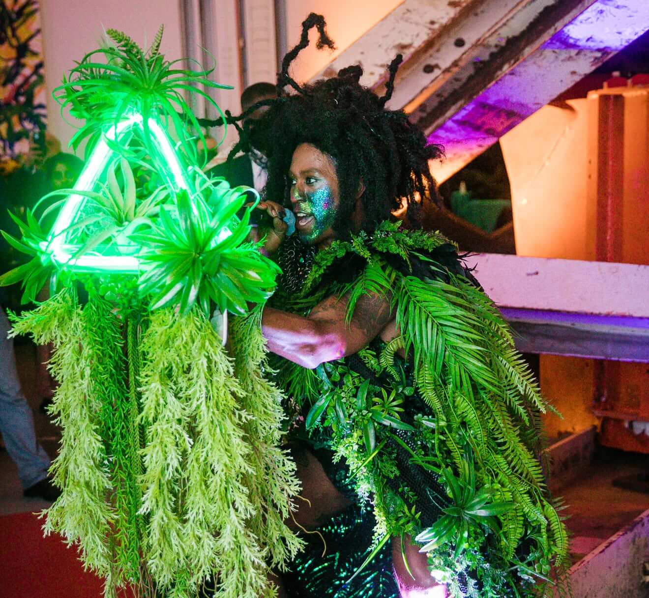 A brown-skinned gender nonconforming person holding a microphone sitting down. This person has black locs and blue and green sparkling makeup on their face, wearing black with green plants draped over their clothing. They are performing in front of a neon green triangle with green flowers and plant vines hanging from it.