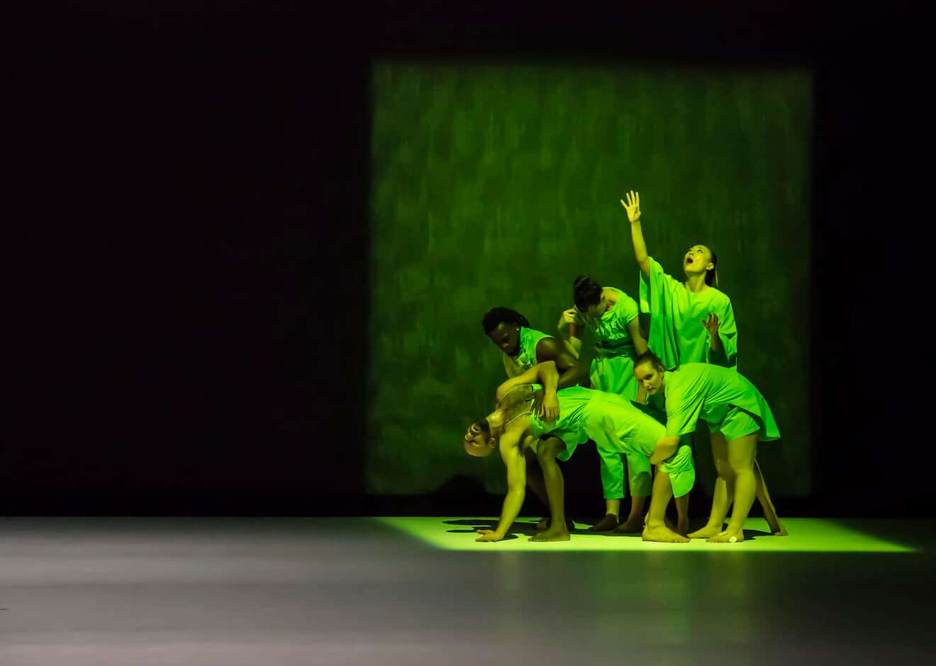 Five dancers in bright green costumes, clumped together, re-enacting a and scene. One figure has fallen backwards, and his body is limp. Another holds the other’s fallen body on his thighs and looks down at him. Two women are standing up, one has her hand curled onto her forward in a gesture of grief, the other looks up with her hands open and lifted and her eyes rolling upwards. The fifth figure crouches, holding the shins of the fallen figure, while turning her head directly toward the camera.