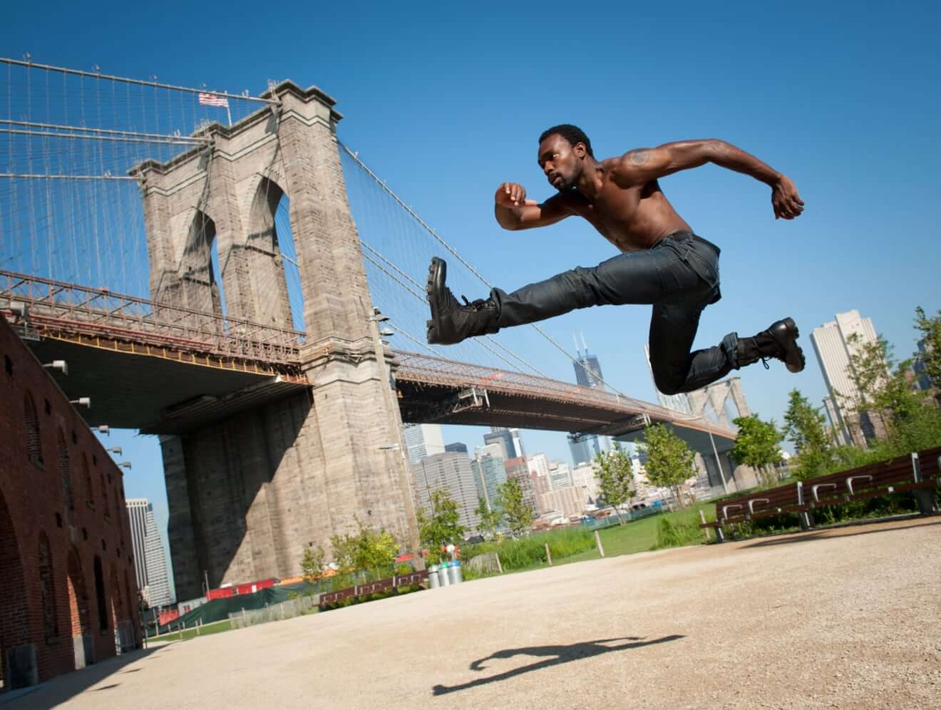 A dark-skinned man wearing jeans and black boots in a hurdle jump towards the Brooklyn Bridge with the backdrop of the Manhattan skyline and blue sky.