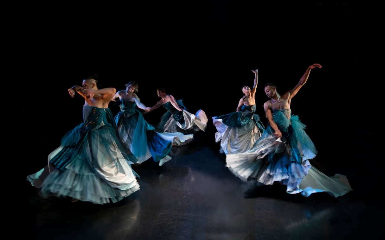 Five gender-nonconforming, trans, and queer dancers on stage. They are all wearing elaborate floor-length blue and silver strapless ball gowns and are dancing in a circle. As the dancers spin around, the skirts of their gowns are flying out with their momentum; each dancer is stretching out their arms in different expansive positions.