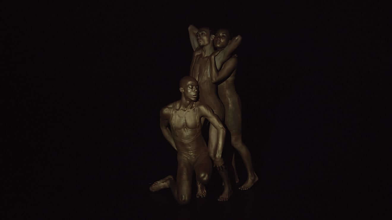 Three Black dancers are in formation glittering in gold body paint. Most forward is a Black man whose right knee is on the ground, his head is turned to the left and both his hands are touching the dancer behind him. The second dancer is standing upright with his back slightly arched and arms over his head connecting with the dancer behind him. The final dancer is also standing and has her arms around the second dancer’s shoulders with her face pressed next to his.