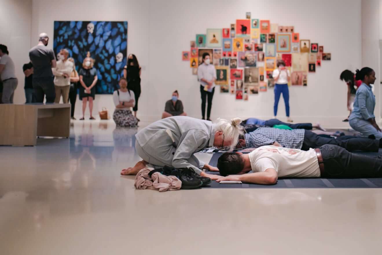 Artist Yanira Castro, a light-skinned Puerto Rican woman with white hair pulled back, kneels over a person who is lying face down on a black floor. Other audience members are laying down in rows behind and next to this person. An audience observes standing and sitting in a white gallery.