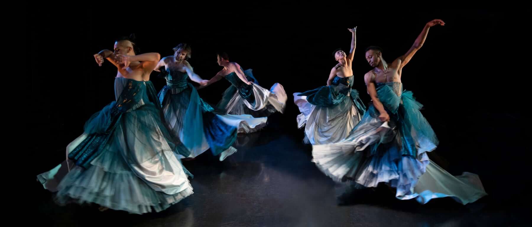 Five gender-nonconforming, trans, and queer dancers on stage. They are all wearing elaborate floor-length blue and silver strapless ball gowns and are dancing in a circle. As the dancers spin around, the skirts of their gowns are flying out with their momentum; each dancer is stretching out their arms in different expansive positions.