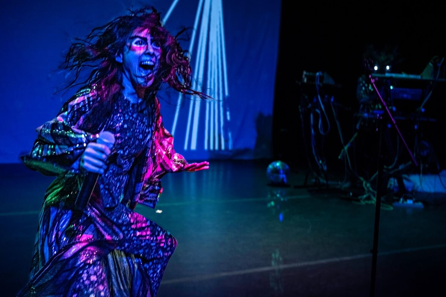 A person wearing a patterned bodysuit and dark reflective jacket crouches in the left foreground and holds a microphone in their right hand. They appear to be yelling or singing, and their long black hair is swinging out as if they've been photographed midway through a large physical movement. They are lit in red and purple. In the far background there is a blue backdrop upon which are projected white stripes that converge at a point just outside the boundaries of the photograph. The stage is otherwise dark, and on the right side in the middle distance you can faintly make out a DJ table with cables and equipment.
