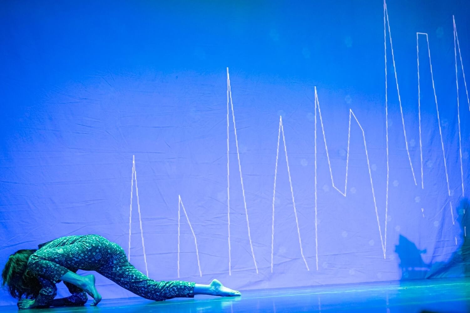 A figure in a patterned bodysuit moves across the stage from right to left in a stylized and elongated crawling pose, with their body and face close to the stage. They are lit in blue and green lights, and the blue backdrop behind them is illuminated with a brighter light, upon which is projected in thin white lines what look like the jagged spikes of a graph or ongoing measurement similar to an EKG.