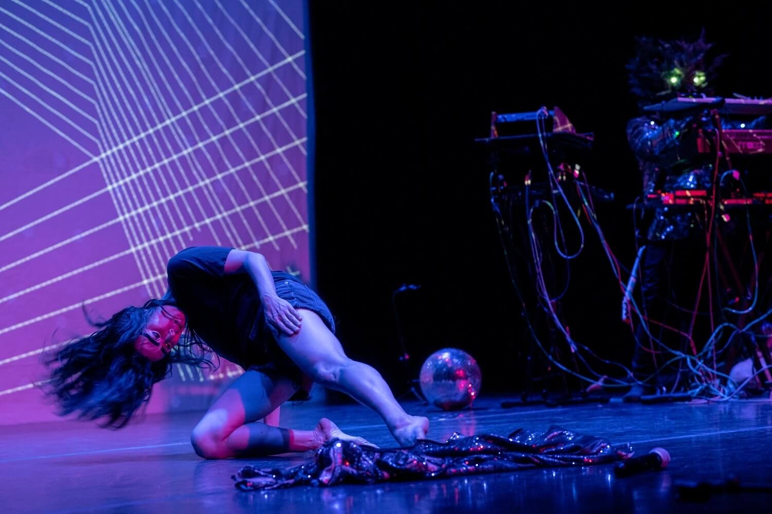 On a stage lit in purple and blue, a female-presenting dancer in a short-sleeved dark shirt and dark shorts presses a knee onto the ground and extends the other leg, while tilting her head down and swinging it so that her long black hair flies out away from her skull.  In front of her is a rumpled and highly reflective swatch of fabric, and behind her a disco ball rests on the ground. The left side of the background shows part of a backdrop for the performance, which is a series of sets of thin white lines crossing over each other like string art. On the right side we can see part of what appears to be a DJ table with cables and equipment. 