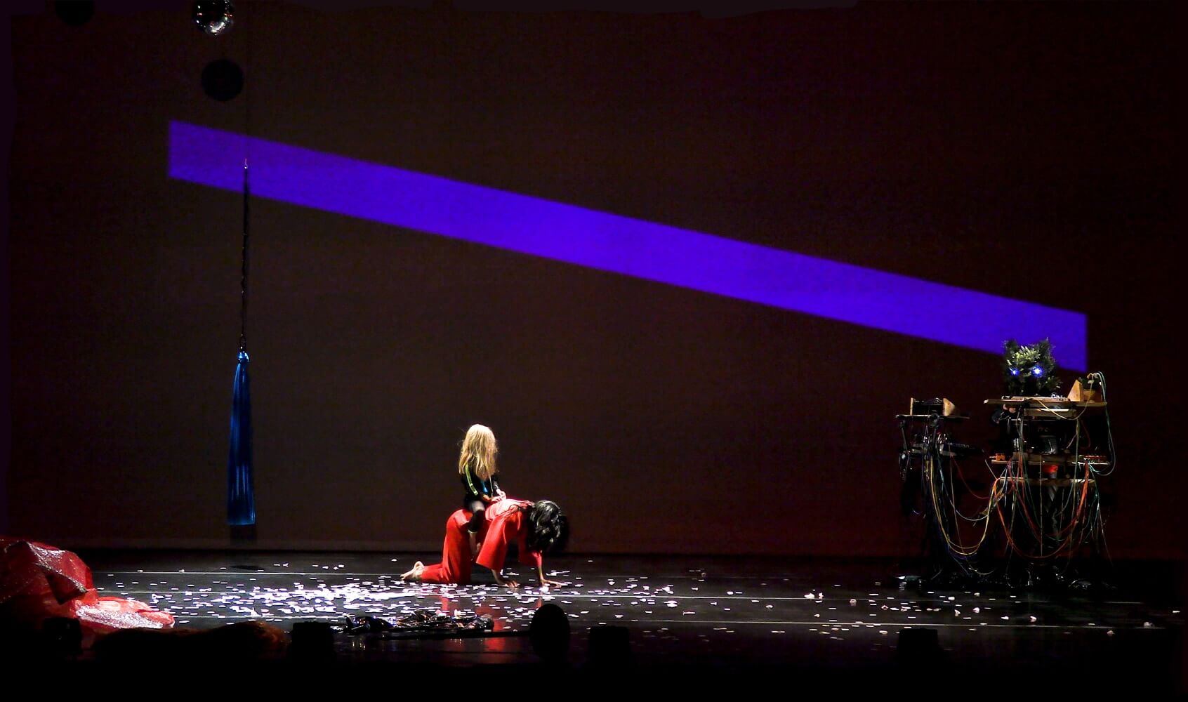 On a darkened stage covered with what appear to be ripped pieces of paper, a figure in a red pant and matching red shirt, and wearing a black helmet the obscures their face, crawls on their hands and knees. A smaller figure dressed in black with long yellow hair sits on the crawling figure, as if riding them.  A thick, bluish-purple diagonal stripe runs across the backdrop from the upper left to the bottom right. On the far right side of the stage is a DJ table with cables and equipment.