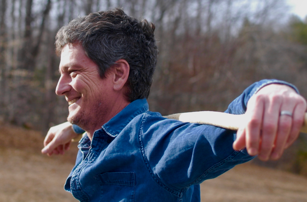 Close up profile photo of a man standing in a field and grinning. He has short, dark brown hair that's peppered with gray, and he's wearing a dark blue denim long-sleeved shirt. His arms are extended and draped around a long wooden pole that runs behind his neck. Photo by Skye Simonet.