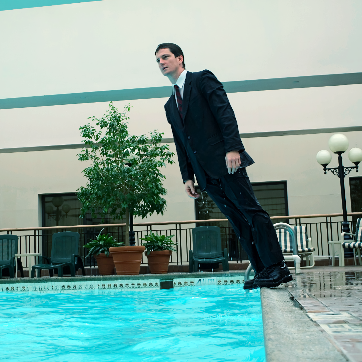 A man with short dark hair, wearing a dark, dripping wet suit and tie, stands in a relaxed pose and looks into the distance as he falls into a large pool.